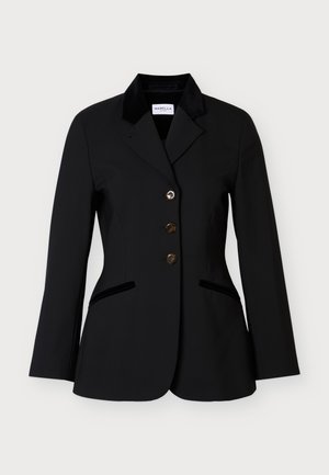 Femme debout, portant un blazer noir cintré à trois boutons, un pantalon large assorti et des chaussures noires pointues.