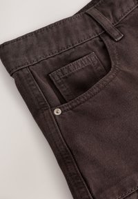 Jeans in denim marrone scuro con tasca frontale arricchita da dettagli di cuciture e bottone decorativo. Il tessuto ha una consistenza liscia.