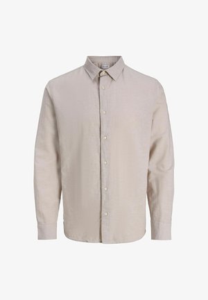 Jeune personne aux cheveux bouclés portant une chemise beige boutonnée sur un tee-shirt graphique noir, un jean large noir et des baskets blanches, regardant vers la droite.