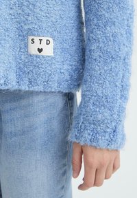 Mėlynas tekstūruotas megztinio rankovės ir apdailos kraštas su baltu pleistru, pažymėtu "STD" ir širdimi, derinamas su šviesiai mėlynomis džinsinėmis kelnėmis ir atsipalaidavusia ranka.