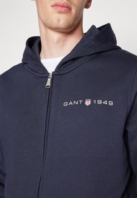 Felpa con zip di colore blu navy, con un logo a contrasto "GANT 1949" ricamato sul petto, dotata di un cappuccio foderato e una texture di tessuto liscia.