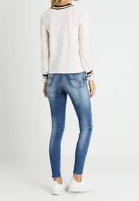 Leichtes, langärmliges weißes Oberteil mit einem schwarz-gelb gestreiften Kragen, kombiniert mit blauen Skinny-Jeans und weißen Sneakers, von hinten betrachtet.