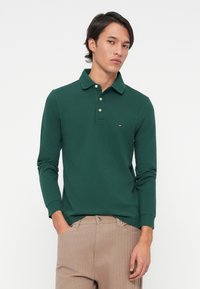 Jonge man met kort donker haar draagt een donkergroene polo met lange mouwen en beige broek, staand met één hand in zijn zak tegen een witte achtergrond.