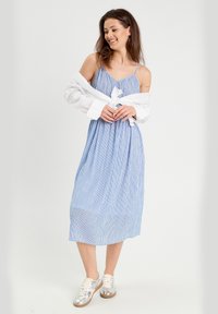 Robe maxi à rayures bleues et blanches avec des fines bretelles, taille cintrée et jupe fluide. Associée à des baskets argentées et une chemise blanche.