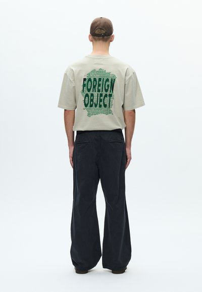 Μπεζ υπερμεγέθη μπλούζα με πράσινο κείμενο "FOREIGN OBJECT" στην πλάτη, συνδυασμένη με χαλαρά σκούρα παντελόνια και καφέ καπέλο.
