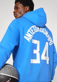 Mėlyna gobtuvas su kapišonu, ant nugaros yra balti užrašai "ANTETOKOUNMPO" ir numeris "34". Užrašai turi perforuotą raštą. Šalia yra krepšinis.