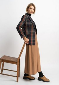 Schwarze und rote karierte Blazer mit strukturierten Schultern über einem beigen Maxirock. Schlanke schwarze Socken und braune klobige Loafers runden das Outfit ab.