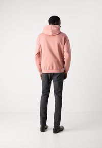 Sudadera con capucha rosa de corte relajado, con dobladillo y puños de canalé, combinada con jeans gris oscuro y zapatos negros, visible desde la parte trasera.