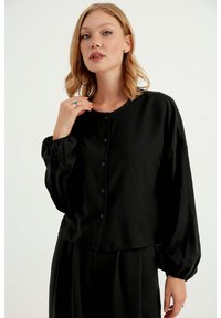 Blusa nera con bottoni, maniche lunghe e a sbuffo, tessuto morbido, scollatura tonda e taglio leggermente cropped. Dettaglio di trama sottile visibile.