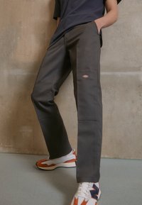 Personne portant un pantalon de travail gris foncé avec le logo Dickies, un t-shirt noir et des baskets New Balance blanches et orange, debout contre un mur en béton.