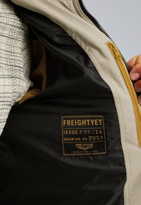 Intérieur d'une veste beige avec une doublure noire et une fermeture éclair jaune. Une étiquette affiche "FREIGHTYET" avec des spécifications et "testé en haute altitude".