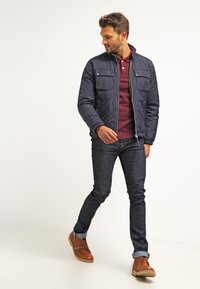 Chaqueta acolchada navy con dos bolsillos frontales, combinada con un polo burdeos, jeans oscuros y zapatos marrones. Enfocándose en la textura y el diseño en capas.