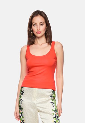 Frau trägt ein ärmelloses, leuchtend orangefarbenes Oberteil und helle Hosen mit grünen und schwarzen Blumenmustern und steht vor einem schlichten Hintergrund.