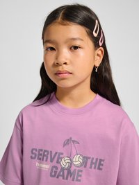 Junges Mädchen mit langen dunklen Haaren, rosa Haarklammern, trägt ein pinkes "Serve the Game" T-Shirt mit Volleyball-Grafik.