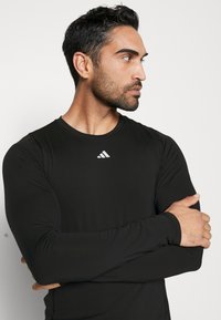 Langärmliges schwarzes Sportshirt aus dehnbarem Material, mit einem kleinen weißen Adidas-Logo auf der Vorderseite. Glatte Textur und taillierte Passform.