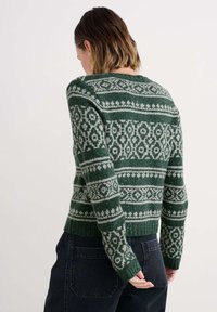 Rövid hajú nő zöld és fehér mintás kötött pulóvert és sötét farmert visel, kezét a háta mögött tartja, háttal áll.