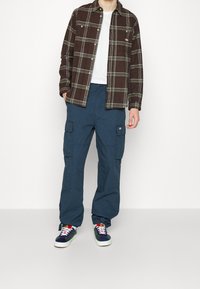 Brun och grön rutad knäppskjorta över en vit t-shirt, kombinerad med blå cargobrallor och mångfärgade sneakers.