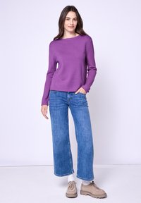 Paarse gebreide trui met een ronde halslijn, gecombineerd met brede blauwe jeans. De jeans hebben een vervaagd textuur en manchetjes bij de enkels.