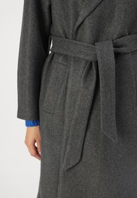 Grijze wollen jas met een ceintuur, grote revers, zijzakken en een gestructureerde stof. De manchetten hebben een blauw gebreid detail.