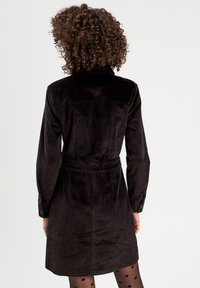 Robe en velours côtelé noir avec un col de chemise, des manches longues, des poignets à boutons et une ceinture à nouer ; présente des côtes verticales et un ourlet légèrement évasé.