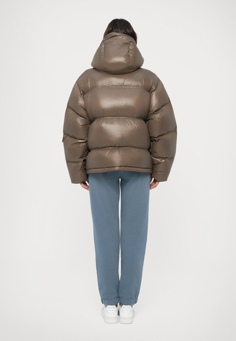 Persoon die met de rug naar de kijker staat, gekleed in een bruine pufferjack met capuchon, blauwe joggingsbroek en witte sneakers, op een witte achtergrond.
