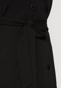 Schwarzer, doppelt geknöpfter Mantel aus strukturiertem Stoff, mit Bindegürtel und großen schwarzen Knöpfen. Fokus auf schlankem Design und utilitaristischen Details.