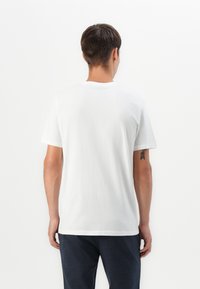 Mand med kort mørkt hår, der står med ryggen til, iført en ensfarvet hvid T-shirt og mørke bukser foran en hvid baggrund.