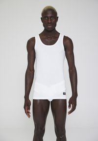 White cotton tank top with a round neckline and a fitted design. The fabric is smooth, and a label is positioned at the bottom left.