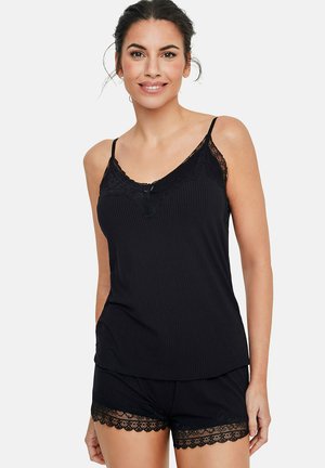 Femme souriante portant un caraco côtelé noir et un ensemble pyjama short assorti avec bordure en dentelle, debout devant un fond uni.