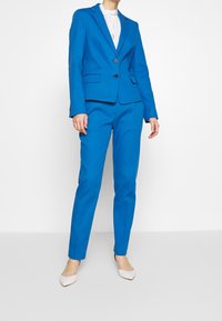 Een persoon staat in een helderblauw op maat gemaakt kostuum met een witte blouse, gecombineerd met lichte hakken, tegen een effen achtergrond.
