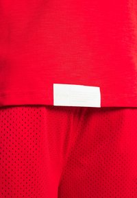 Tela roja con una etiqueta rectangular blanca visible cosida cerca de una costura, encima de un material de malla roja con pequeñas perforaciones.