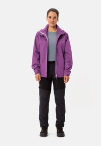 Giacca impermeabile viola con cappuccio, chiusura con zip e strato interno grigio, abbinata a pantaloni da trekking neri e scarpe robuste nere.