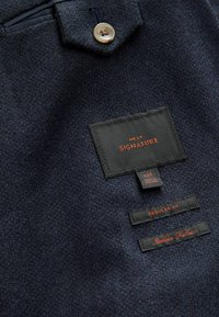 Tissu bleu marine foncé avec un tissage texturé, comportant un bouton, des étiquettes "NEXT SIGNATURE", taille "40R" et "Tissu Italien" en fil orange.