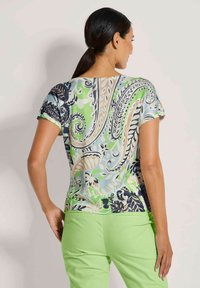 Korte mouwen top met een blauw, groen en beige paisleypatroon; zachte stof met een getailleerde silhoutte. Gepaard met limoen groene broeken.