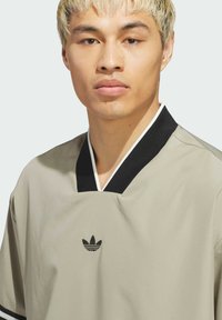 Svetlá šedá tričko s krátkym rukávom a čiernym golierom do V, s malým čiernym logom Adidas trefoil na hrudi. Hladká štruktúra látky.