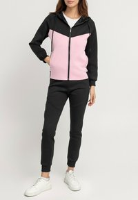 Haut de survêtement zippé noir et rose avec capuche et poches, associé à un pantalon de jogging noir. Des baskets blanches complètent la tenue. Texture de tissu lisse.