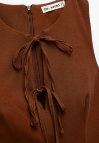 Brązowy bezrękawnik z zamkiem błyskawicznym i dwoma trokami przy dekolcie. Wykonany z gładkiego materiału o subtelnej teksturze. Widoczna etykieta marki.