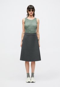 Green textured top with gathered design, paired with a solid dark gray A-line skirt. Model wears light shoes and gray socks, with sunglasses.