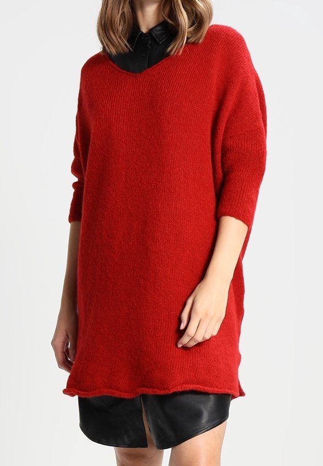 Femme portant un pull en maille rouge ample par-dessus une robe noire à col avec une fente sur le devant, debout devant un fond clair uni.