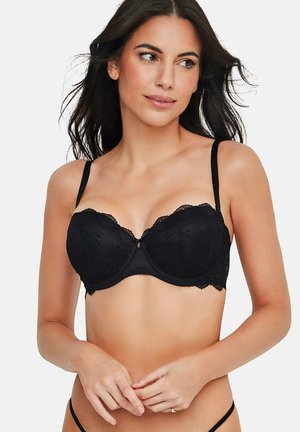 Femme aux cheveux foncés portant un soutien-gorge en dentelle noire, regardant légèrement sur le côté, les mains doucement jointes au niveau de la taille.