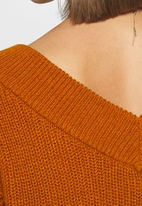 Nahaufnahme von Schulter und Nacken einer Person, die einen orangen Strickpullover und einen goldenen Ohrring mit hängendem rechteckigem Anhänger trägt.