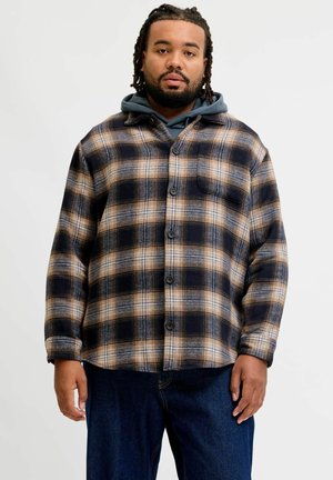 Homme aux cheveux tressés portant un sweat à capuche bleu sous une chemise à carreaux marron et noir boutonnée et un jean bleu foncé, debout devant un fond uni.