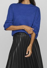 Femme portant un pull bleu à manches longues retroussées et une jupe en cuir plissée noire, tenant doucement son bras.