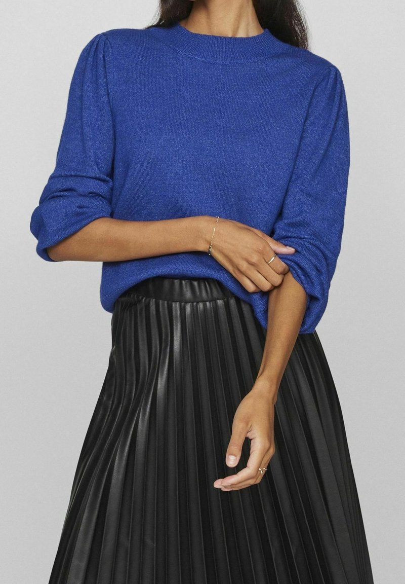 Femme portant un pull bleu à manches longues retroussées et une jupe en cuir plissée noire, tenant doucement son bras.