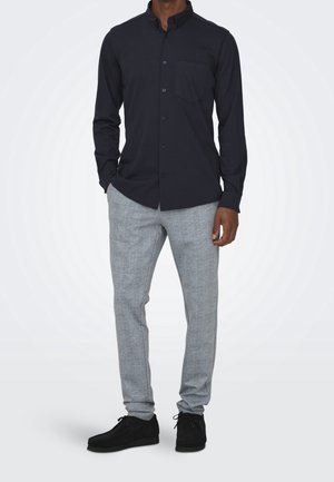 Marineblauwe overhemd met kraag en een borstzak, gecombineerd met grijze gestructureerde broeken en zwarte schoenen, staande voor een effen achtergrond.