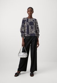 Blouse à motif marine avec des manches bouffantes, associée à un large pantalon noir et un sac à main métallique argenté. Sandales à talons ouverts noires portées.