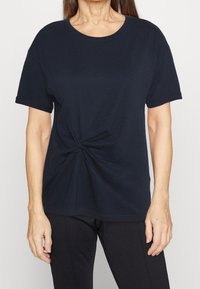 Žena v tmavě modrém tričku s detailem uzlu v pase stojí na světlem pozadí, její ruce jsou odpočívající po stranách.