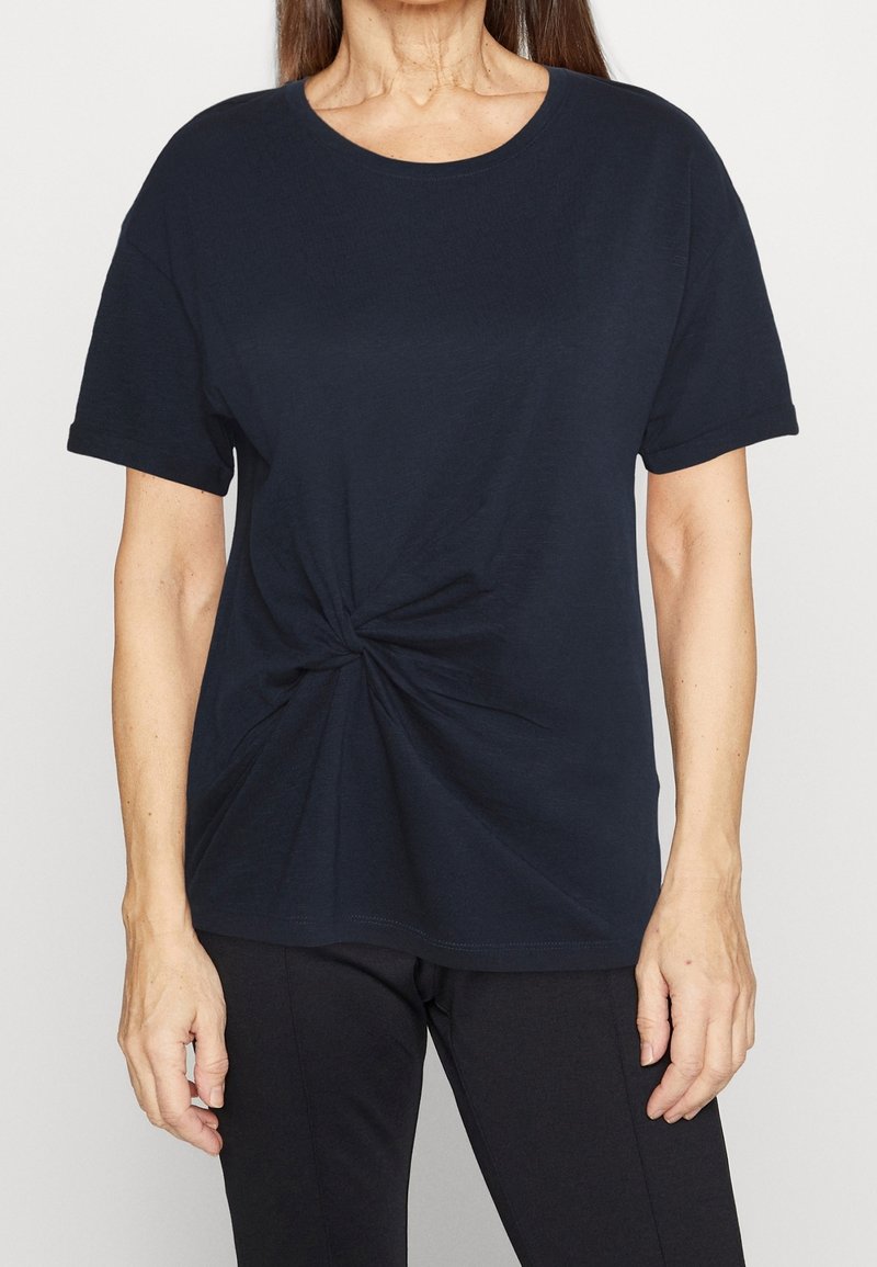 Žena v tmavě modrém tričku s detailem uzlu v pase stojí na světlem pozadí, její ruce jsou odpočívající po stranách.