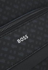 Sac en tissu noir texturé avec une fermeture éclair argentée, arborant un logo "BOSS" blanc en évidence sur le devant.