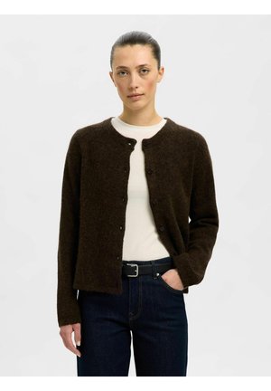 Frau mit zurückgekämmtem Haar, die einen dunkelbraunen, geknöpften Cardigan über einem weißen Top, dunkle Jeans und einen schwarzen Gürtel trägt, steht vor weißem Hintergrund.